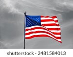 National flag of the United States, American or U.S. flag also called  “Stars and Stripes“, “The Star-Spangled Banner. Colorful fabric waving in the wind on a pole with grayscale sky background.