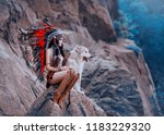 Native American Indian woman. The huntress sits on the rock with the wolf and looks into the distance. Dog Alaskan Malamute. On the girl beads, leather skirt, ethnic jewelry.