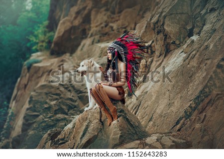 Native American Indian woman. The huntress sits on the rock with the wolf and looks into the distance. Dog Alaskan Malamute. On the girl beads, leather skirt, ethnic jewelry.