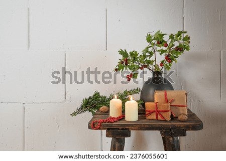 Natural Advent and Christmas decoration, gifts, candles and branches with red fruits in a vase on a rustic wooden stool in front of a rough white wall, copy space, selected focus