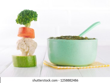 Natural Baby Food Concept. Vegetable Puree In Bowl Near Pyramid Of Brocolli, Carrot, Cauliflower And Zucchini Pieces On White Background With Blank Space For Text.