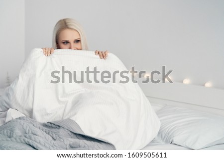 Natural beautiful woman sitting in bed. Girl hiding behind a blanket. Attractive young woman covering half of her face with blanket and looking at camera while sitting in bed at home