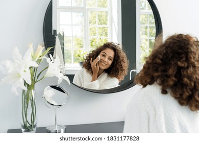 Natural Beauty Concept. Glad Young Afro American Woman Making Morning Routine, Removing Makeup, Looking At Mirror, Holding Cotton Pad In Hands Near Face And Sitting In Bathrobe At Bathroom