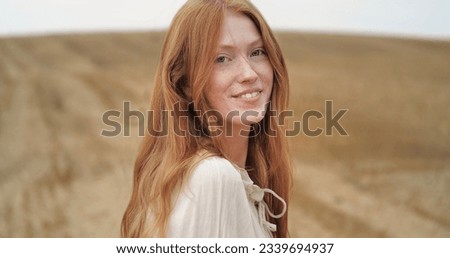 Natural Beauty Ginger Woman is standing on golden Field happily, enjoying Nature. Amazing Woman with long Red Hair, smiling Charmingly. Looking Happy, feeling Liberty at Countryside. Emotions.