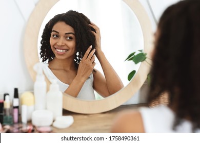 Natural Beauty. Happy Black Girl Touching Her Soft Curly Hair Near Mirror At Home, Enjoying Reasult Of Treatment, Over Shoulder View
