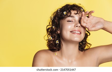 Natural beauty and skincare. Portrait of happy woman with curly hair and healthy face without acne or makeup, chamomile in hair, touching skin and smiling at camera, yellow background - Shutterstock ID 2147051447