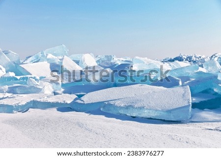 Natural cold background with huge blocks of blue transparent ice floes on snowy hummock field of frozen Baikal Lake on sunny frosty January day. Scenic winter landscape. Ice travel and outdoors