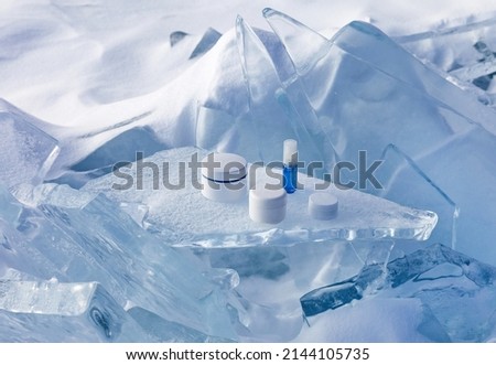 Natural cosmetic products for protecting and nourishing skin of face, lips and hands in cold season. Advertising of cosmetics on blue and white podium made of piece of ice on snowy background