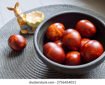 Natural dyed Easter eggs in warm brown and reddish tones arranged in a ceramic bowl on a textured placemat. Traditional holiday decoration with a small bunny figurine holding a shell with salt in the background. Cozy home setting with soft lighting, celebrating spring and Easter traditions