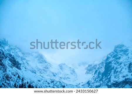 A natural landscape of a snowy mountain with rocks and trees on a cloudy day, showcasing geological phenomenon with a freezing ice cap and cumulus clouds in electric blue sky. Morskie Oko, Poland