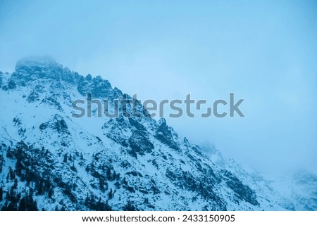 A natural landscape of a snowy mountain with rocks and trees on a cloudy day, showcasing geological phenomenon with a freezing ice cap and cumulus clouds in electric blue sky. Morskie Oko, Poland