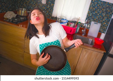 natural lifestyle portrait of young beautiful and happy Asian Korean woman in kitchen apron playing air guitar with cooking pan having fun enjoying domestic chores dancing and singing at home