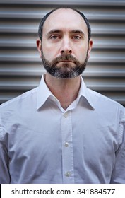 Natural Light Headshot Of A Male. He Has A Neatly Trimmed Beard And Is Wearing A White Shirt And Is Infront Of A Metal Panel. 