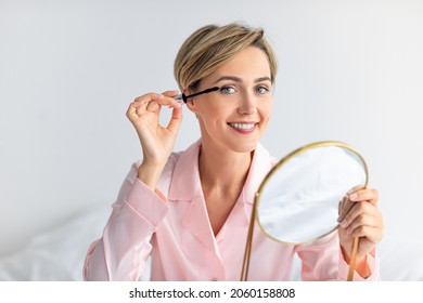 Natural Makeup Concept. Attractive Lady Applying Mascara On Eyelashes Enjoying Morning Beauty Routine Holding Mirror And Looking At Camera. Female Cosmetics Product, Free Copy Space