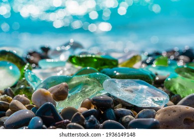 Natural polish textured sea glass and stones on the seashore. Azure clear sea water with waves. Green, blue shiny glass with multi-colored sea pebbles close-up. Beach summer background. 