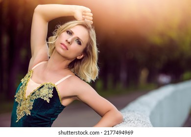 Natural Portrait Of Shy Winsome Professional Caucasian Ballet Dancer In Green Tutu Dress Posing Near Stone Fence In Summer Sunny Park Outdoors. Horizontal Composition