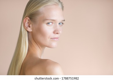 Natural Portrait Of Topless Female Model Looking Over Shoulder Staring Into The Camera With Beautiful Eyes. Isolated Against A Rose Gold Background. Face Beauty, Hair And Skin Care Cosmetics.