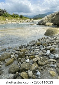A natural river setting captured on surrounded by large rocks and distant green hills or mountains, under a partly cloudy sky. The atmosphere feels calm, fresh and untouched
