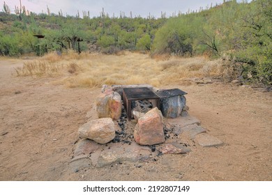 Natural Rock Fire Pit With Box Grills In The Middle At Tucson, Arizona. Fire Pit On A Dry Dirt Ground Near The Wild Shrubs And Saguaro Cactuses On A Slope At The Background.