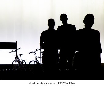 Natural Silhouettes Of A Young Woman Walking And Two Young Men Looking At Her From Behind, In Front Of The Modern Building In The Night