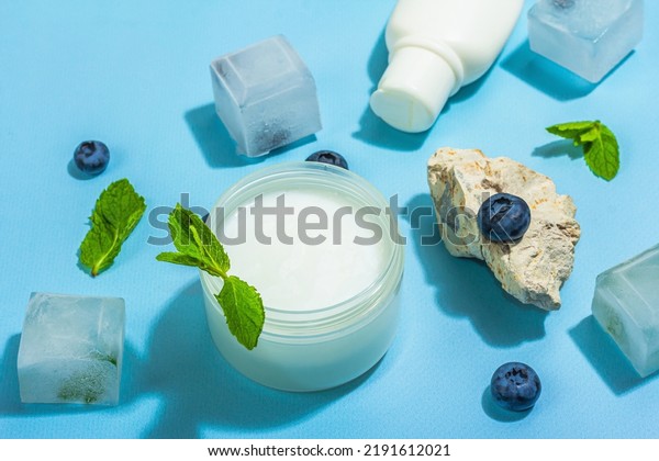 Natural skincare. Cream or lotion with organic\
berries. Blueberry, mint, and ice. Moisturizing and tonic cosmetics\
concept, flat lay, trendy hard light, dark shadow, blue background,\
close up
