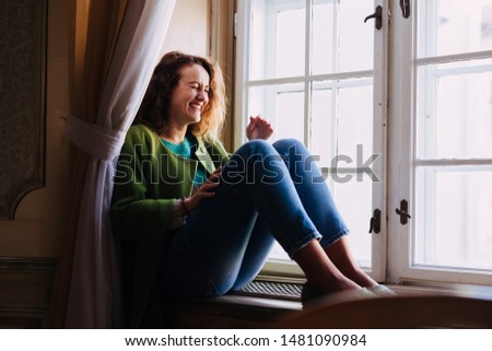 Natural woman laughing spontaneously  near a window. Warm dressed caucasian girl with curly hair.