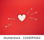 Natural wooden heart shape is targeted by arrows on bold red background. Minimal ready for romance concept with copy space. Flat lay Cupid love idea. Valentines day Holiday.