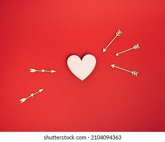 Natural wooden heart shape is targeted by arrows on bold red background. Minimal ready for romance concept with copy space. Flat lay Cupid love idea. Valentines day Holiday.