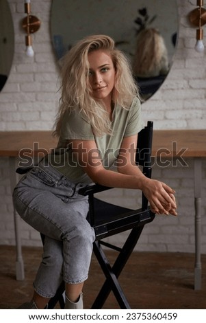 Natural-hued portrait of a blonde beauty with voluminous hair, gracefully seated on a chair in a makeup artist's room