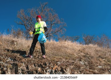 Nature Cleaner Volunteer Activist In Yellow Vest Puts Glass Bottle In The Blue Trash Bag Outdoors