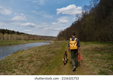 Nature Of The Moscow Region, The Moskva River In The Countryside On A Warm Summer Sunny Day. Man Wears Backpack Walks With German Shepherd Along Path River. Owner And Dog In Park, Rear View.