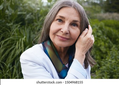 Nature, Relaxation, People And Mature Age. Close Up Picture Of Stylish Friendly Looking Caucasian Sixty Year Old Female Posing Against Green Plants Background, Smiling, Touching Her Loose Gray Hair