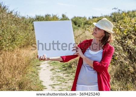 nature teasing - relaxed 50s woman with summer hat showing a white blank cutout to write message on,walking on countryside path

