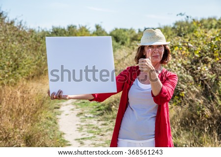 nature teasing - smiling 50s woman with summer hat agreeing to message on white blank panel,walking on countryside path

