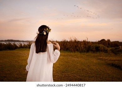 Nature, woman and back with flower crown at sunset for summer, scenery and peace in meadow. Blue sky, clouds and birds by person with floral accessory for purity, salvation and renewal in faith - Powered by Shutterstock