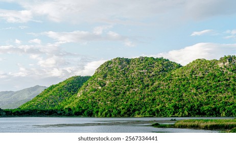 Nature's Reflection. A verdant mountain range slopes down to a tranquil lake, its reflection shimmering on the placid water. Lush greenery covers the hills, creating a serene and picturesque landscape - Powered by Shutterstock