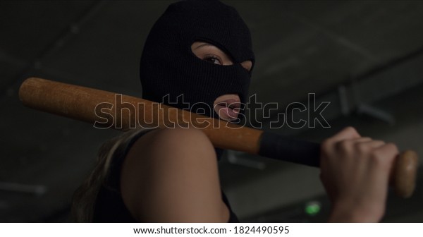 Naughty in a balaclava holds a\
baseball bat in her hands and poses on the territory of a car\
parking. Cheeky sexy woman in mask shows disrespect and\
aggression