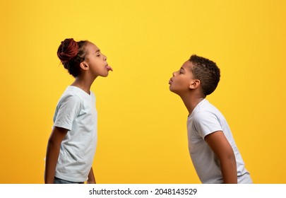 Naughty black brother and sister having fight, afro-american boy and girl showing each other tongues, yellow studio background. Siblings relationships, brotherhood, sisterhood, kids quarrels concept