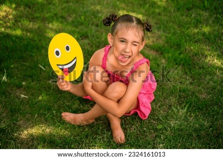 Naughty funny crazy girl makes a grimace and shows her tongue laughing merrily and holding a smiley face in her hands.