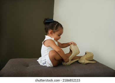 Naughty happy little girl putting on and playing with her mom's heels at home making mischief sitting in an armchair
