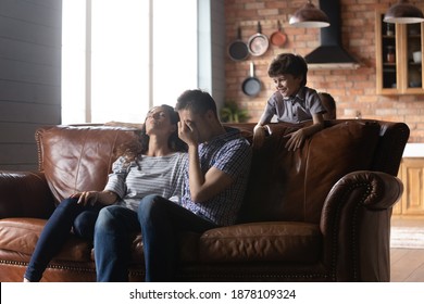 Naughty Kids. Stressed Millennial Couple Parents Of Two Little Children Sit On Couch At Home Feeling Tired Of Noise Mess. Exhausted Mom Dad Resting On Sofa While Small Active Son Daughter Run Around