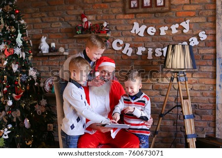 Naughty male children of European appearance want Christmas grandfather to show them interesting cartoons on gadget in decorated room with high floor lamp, walls which posters hang, and tree dressed