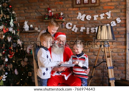 Naughty male children of European appearance want Christmas grandfather to show them interesting cartoons on gadget in decorated room with high floor lamp, walls which posters hang, and tree dressed