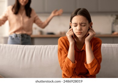 Naughty teen girl covering ears and closing eyes sitting on couch refusing to fulfill mother requests, aggressive woman yells at daughter - Powered by Shutterstock