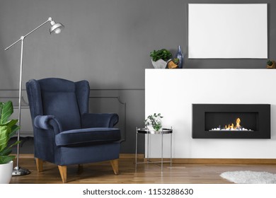 Navy Blue Armchair Between Lamp And Silver Table In Living Room Interior With Fireplace. Real Photo