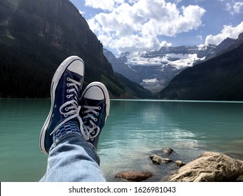 Navy blue Chuck Tylor Converse All-Star sneakers resting rocky shoreline of glacier-fed aqua green Lake Louise with the glacier filled Canadian Rockies, framing a cloud filled blue sky.