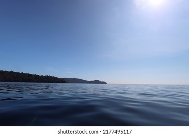 Navy Blue Sea Scene With Silhouette Island Far Away At Horizon With Smooth Blue Sky And Light Of Sun. More Space For Text