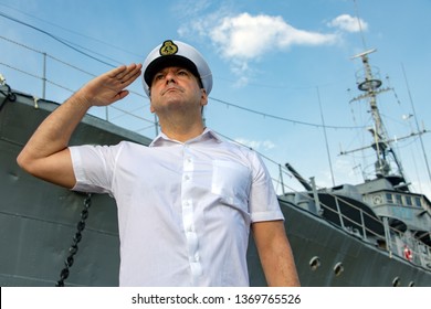 Navy Officer Standing Beside Warship And Do Salute.The Captain In White Uniform Stands Under A Battleship And Saluting.