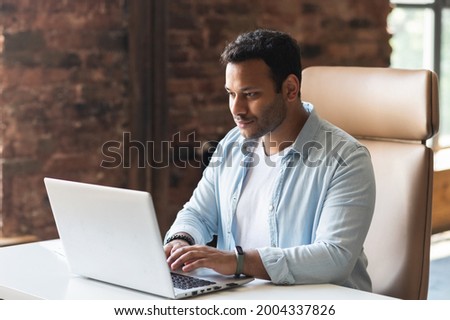 ndian developer guy is working with a laptop sitting at the desk indoors. A young multiracial guy in smart casual wear is typing on the keyboard, answering email, communicates with the customer