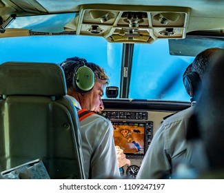 Ndutu, Tanzania, May 3, 2018: Rear View Of Pilot And Co-pilot Sitting In Cockpit Of Light Plane, In Flight. They Are Wearing Earphones. Control Panel Is Showing Map. Prop Is Just Visible. Africa.